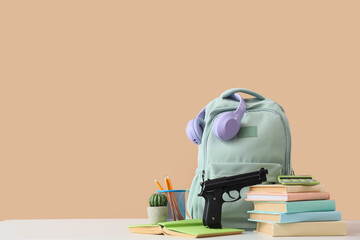 Backpack with gun, headphones and books on table against beige background. School shooting concept