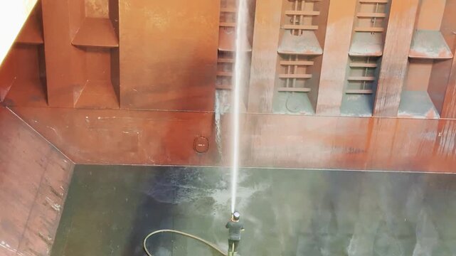 High angle view of manual labor inside a ship's hold: a sailor cleaning dirty steel bulkheads with water spray after cargo discharge.
