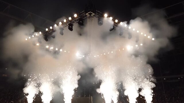 Spectacular stage lighting truss illuminating smoke and white pyrotechnic fountains during a major live arena concert.