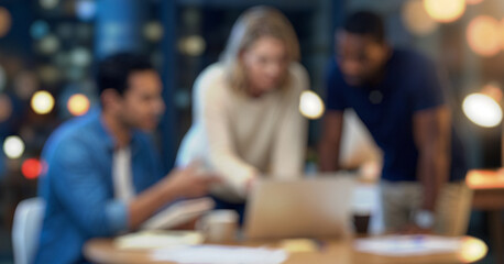 Three individuals engage in a discussion around a laptop in a blurred indoor setting with warm lighting. Papers and coffee cups are also visible.