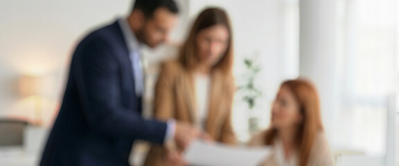 Blurred image of three professionals discussing paperwork in a modern office setting. Two are standing and one is seated, all focused on the document being shared.
