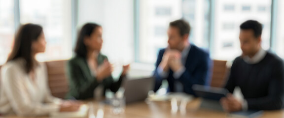 Blurred image of a business meeting with four professionals seated around a table. They appear to be engaged in discussion, sharing ideas in a modern office setting.