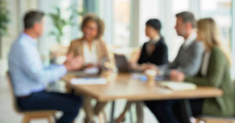 Blurred image of a group meeting in a bright room. Several individuals sit around a table engaged in discussion, with laptops and notebooks visible.