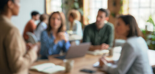 Blurred image of a group in a meeting setting, focusing on a speaker in the foreground. Participants engaged in discussion with laptops and notebooks present.