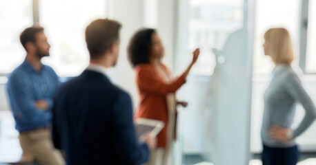 Blurred image of a diverse group of professionals engaged in a meeting. One person is writing on a transparent board while others observe attentively in a bright office.