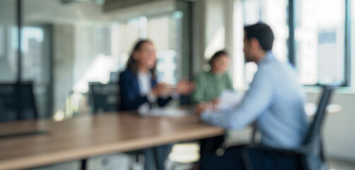 Blurred image of three individuals in a meeting. Two are engaged in discussion while one listens. The setting features a modern office with large windows.