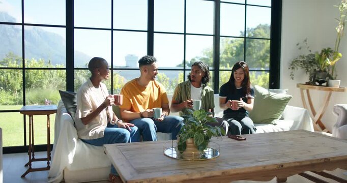 Diverse friends sitting on sofa in living room, exchanging smiles sparking casual chat with mugs