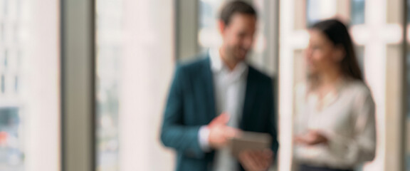 Blurred image depicting two business professionals engaged in conversation while looking at a digital tablet. The setting appears to be a modern office or workspace.