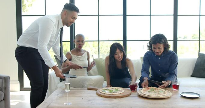 Diverse friends seated on sofa reaching for pizza while standing man in white shirt placing plates