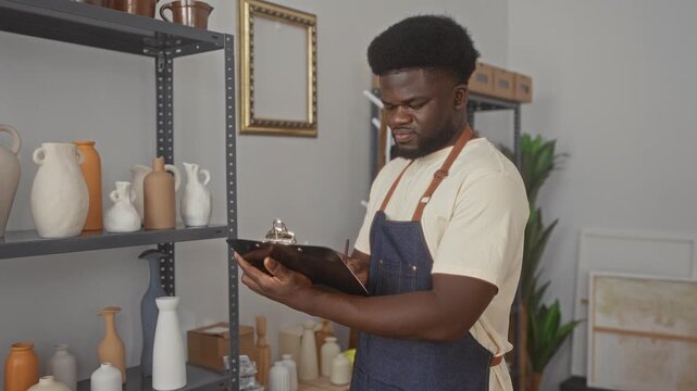 Man in apron holding clipboard and pen writing inventory by shelves of pottery in studio; focus craft precision.