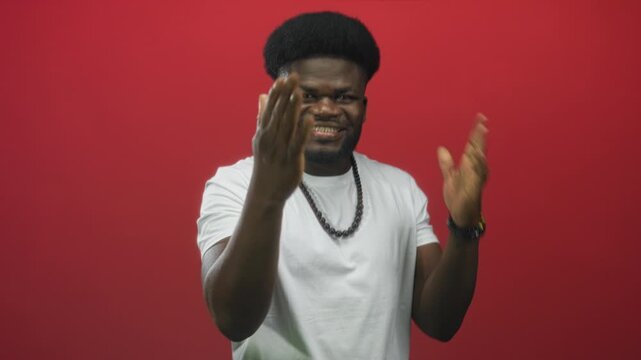 Man extends hand toward camera, smiles and claps while wearing white t shirt and beaded necklace in studio with red wall; joy celebration energy.