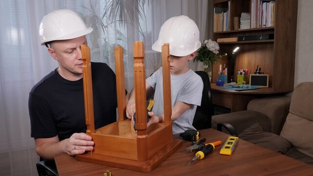 A young lad and his father, both wearing white helmets, are working on a wooden chair at home. The man is using a screwdriver, while the boy is using a level.