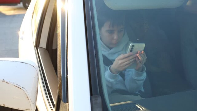 Child seated in the front passenger seat of a car using a smartphone while secured with a seat belt. Filmed through the front windshield with visible reflections and natural light inside the vehicle. 