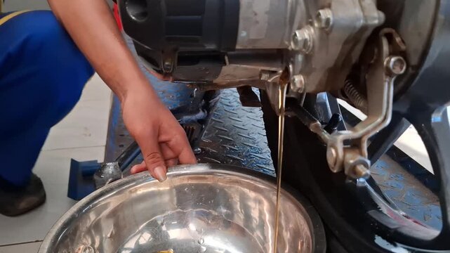 Close-up of a vocational student performing a scooter engine oil drain during motorcycle maintenance training, collecting used lubricant into a metal container in a workshop environment.