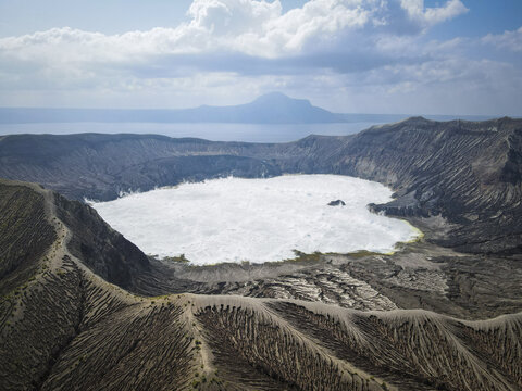 Aerial view of Taal Volcano's crater lake shimmers under the cloudy sky, a stark contrast to the rugged, textured landscape of Taal Volcano Island, Taal Lake, Batangas, Philippines.