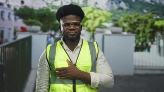 Man wearing neon safety vest and glasses with hand on chest on urban street near white fence and building; pride duty.