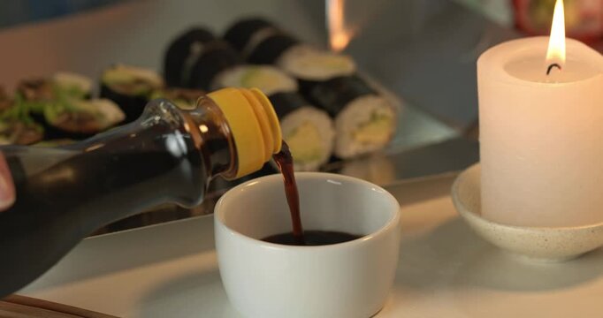 Soy sauce being poured into a bowl on a background of a box of takeaway rolls. Rolls with avocado, leek and shiitake mushrooms. Sushi eating women alone evening in candle light.