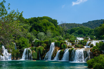 Naklejka premium Stunning series of cascading waterfalls surrounded by lush greenery, with clear blue water flowing over moss-covered rocks under bright sky. Krka National Park is of Croatia's natural treasures.