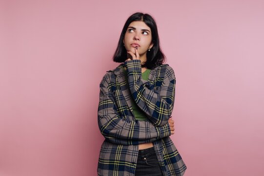 Thoughtful young woman in a plaid shirt poses against a soft pink backdrop, pondering life&rsquo;s questions with a curious expression
