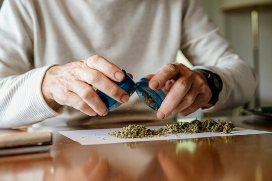 Midsection of elderly man removing weed from grinder on paper at home