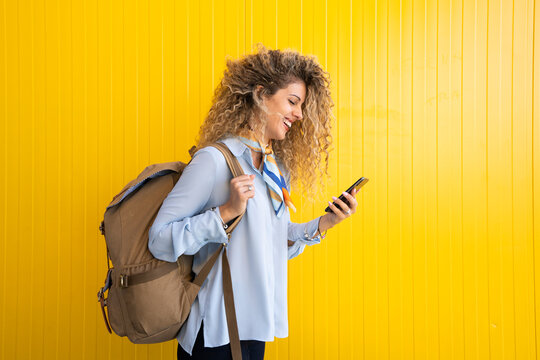 Smiling young woman with backpack in front of yellow background looking at cell phone