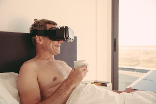 Man wearing VR glasses in bed holding cup of coffee