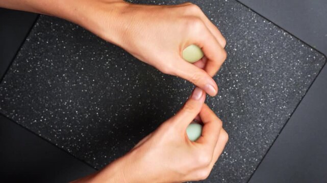 Hands pressing two green objects on a dark surface in a kitchen setting. Two hands press down on green items against a dark kitchen surface.