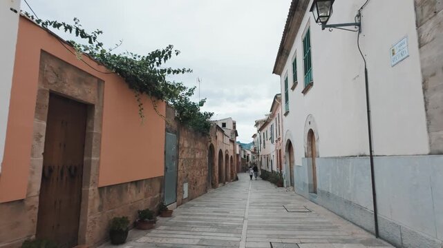 First-person view walking through sun-drenched, narrow Carrer de la Rectoria, observing historic residential architecture in Alc&uacute;dia's ancient walled city, Mallorca, Spain.