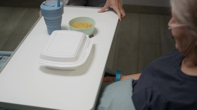 Tracking shot of unrecognizable nurse in white coat bringing meal tray to senior Caucasian female patient lying in bed and feeling grateful for hospital service