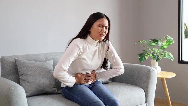 Young woman sitting on couch is experiencing intense abdominal pain. She is holding her belly and grimacing in discomfort