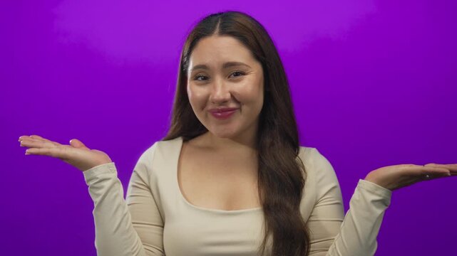 Young hispanic woman shrugs shoulders and holds open hands in purple studio in a questioning gesture; uncertainty.