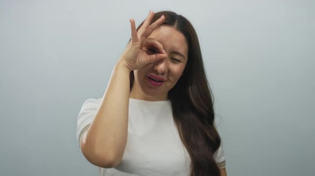 Smiling young hispanic woman shows ok sign in a pale blue studio setting with visible hand gesture; approval confidence.