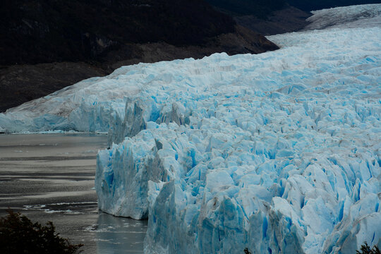 View of the Perito Moreno Glacier's icy blue expanse meets the dark waters below, framed by rugged mountains, Los Glaciares National Park, Santa Cruz Province, Argentina.