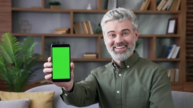 Smiling mature bearded man with gray hair sitting on sofa at home showing smartphone with green screen. Confident male looking camera holding mobile phone with chroma key for app or advertising layout