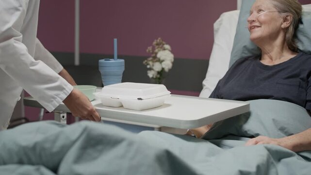 Caring middle-aged Caucasian female doctor in white coat bringing mobile tray table with food to senior female patient lying in hospital bed, serving meal in modern well-equipped ward