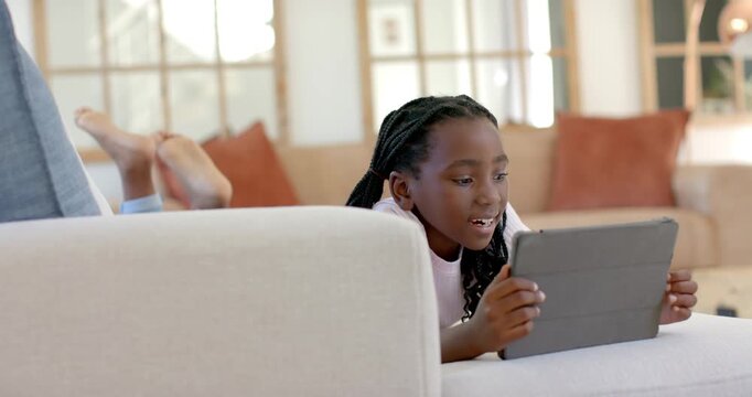 African American child lying on sofa at home holding tablet in folio case, reacting to screen