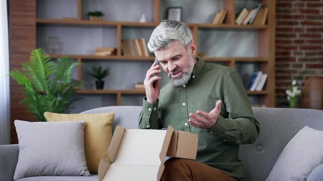 Bearded man with gray hair sitting on sofa at home angrily talking on phone. Frustrated male customer arguing about damaged package, complaining about bad service and leaving negative review online