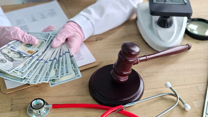 Hands in gloves hold cash near a gavel and stethoscope on a wooden table during a legal or medical...