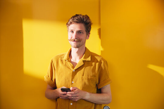 Portrait of smiling male business professional holding smart phone standing near yellow wall