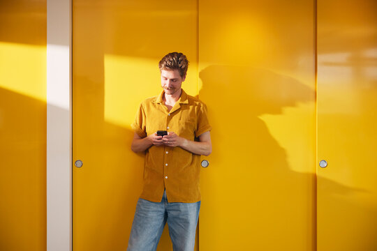 Male business entrepreneur using smart phone while standing near yellow wall in office