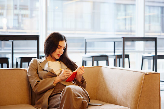Female business professional writing in personal organizer while sitting on sofa at office