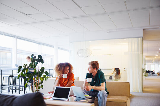 Male and female business colleagues talking with each other during coffee break while sitting on sofa in office