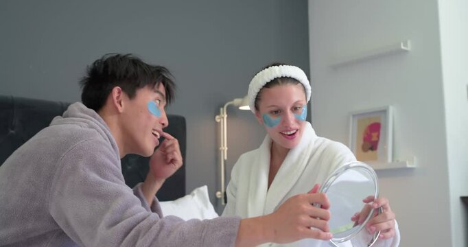 With blue patches Asian man and African woman in bathrobes holding mirror checking patch fit