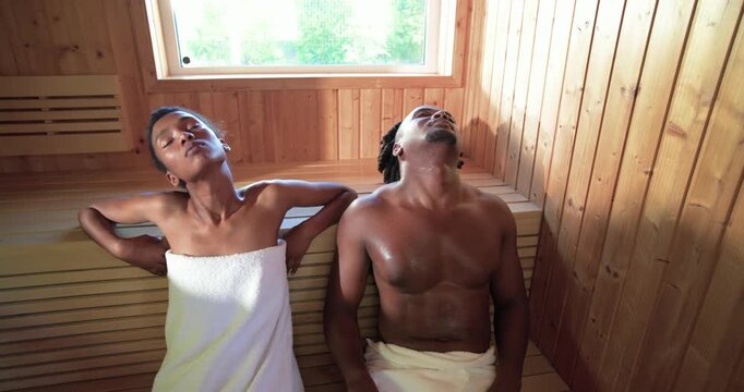 African American couple sitting on bench under pane, sauna heat causing leaning with towels, heater