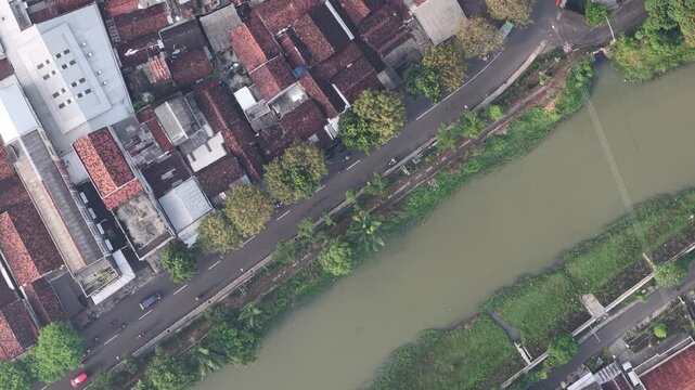 Bird's-eye view of a muddy river running between tree-lined banks and dense rows of red-roofed houses, with streets and lush greenery creating a calm, urban-rural contrast.