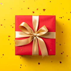 Red gift box with gold ribbon on a bright yellow surface scattered with small red star-shaped confetti