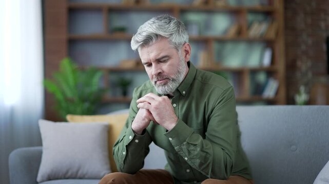 Close-up of mature bearded man with gray hair sitting alone on sofa at home looking sad and depressed. Thoughtful male suffering from stress, loneliness and emotional pain in cozy living room interior
