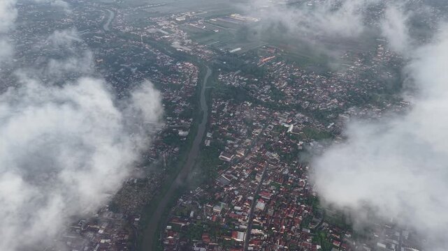 Aerial panorama of a dense town beside a winding river, partly shrouded by clouds. Showcases rooftops, streets, and surrounding fields, conveying urban life, travel routes, and natural contrast.