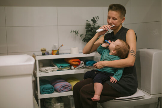 Mother brushing teeth while sitting with daughter on toilet seat in bathroom