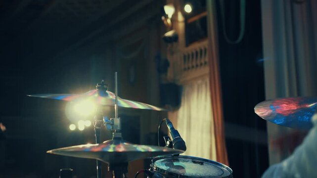 A drummer plays his drums during a concert, with a spotlight shining on him. Dust particles can be seen flying in the bright light above the hi-hat, adding to the atmosphere of the performance.